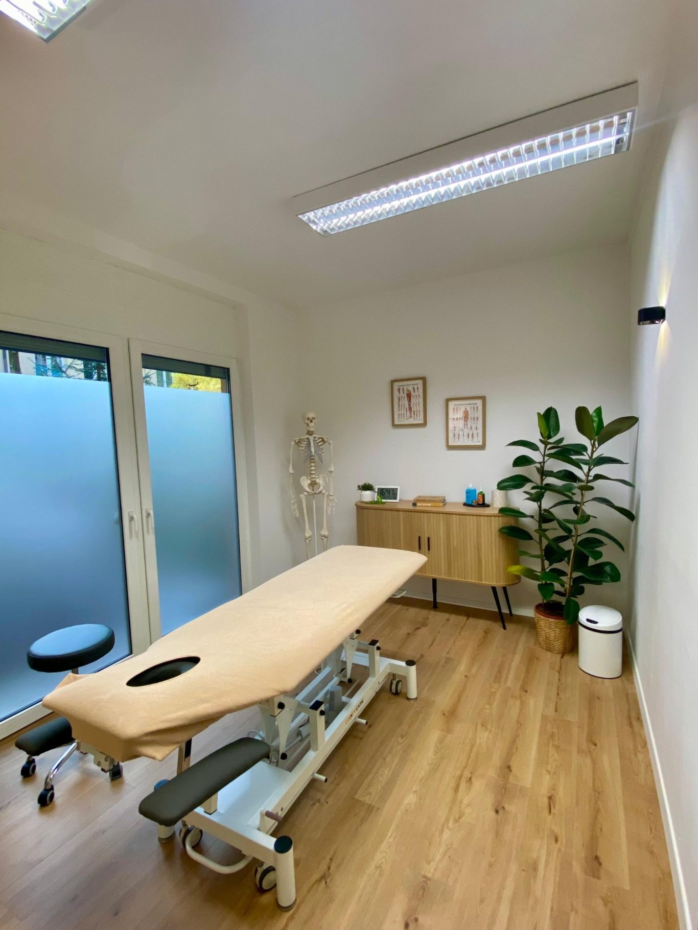 Direkter Blick in den Behandlungsraum auf die Behandlungsliege und die dahinter befindlichen Fenster mit Zugang zum ruhigen Innenhof.